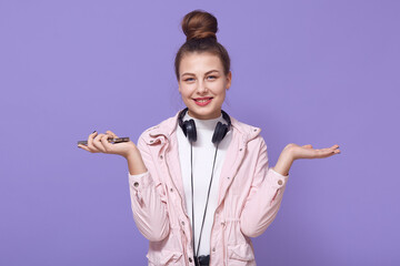 Winsome young European girl in pale pink jacket stands on lilac background with headphones on her neck and spreads hands with the phone aside, looks at camera with charming smile.