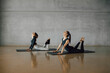 © BASILICOSTUDIO STOCK - Brunette female coach mentoring a woman to practice Pilates stretching exercise, workout during personal Yoga class in a big fitness center with gray concrete wall background. Health club.
