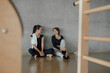 © BASILICOSTUDIO STOCK - Portrait of two sporty beautiful young women in sportswear sitting beside concrete wall of a Pilates center, gym, talking together. Candid funny fitness instructors waiting for class to start.