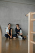 © BASILICOSTUDIO STOCK - Portrait of two sportive beautiful young women in sportswear sitting beside concrete wall of a industrial Pilates center, looking at camera smiling. Female friends waiting for class to start.