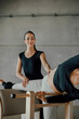 © BASILICOSTUDIO STOCK - Smiling brunette female coach helping a woman to practice Pilates stretching exercise in a ladder barrel equipment, workout during personal class in a fitness center gym, health club.