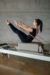 © BASILICOSTUDIO STOCK - Full shot of a strong body of a Pilates Instructor in V sit on a bed of a Reformer machine, pulling a stretch band with hands, training in a big Industrial Fitness Center Studio. Full length.
