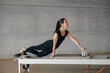 © BASILICOSTUDIO STOCK - Strong Beautiful Fitness young woman in Athletic Black Workout Clothes Doing Stretching Pilates Exercise on a Reformer Machine Bed. Training at Gym, big industrial loft studio.