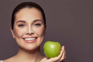 Wall Mural - Beautiful woman with cute smile with white teeth smiling and holding green apple on brown background