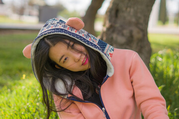 sweet Asian woman in hoodie making faces - lifestyle portrait of young happy and beautiful Korean girl sitting playful on green grass in city park smiling cheerful having fun