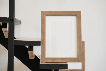 Closeup of square empty wooden picture frame on wooden stairs. White wall background. Scandinavian interior, home design. Art concept. Artistic mockup scene. Front view