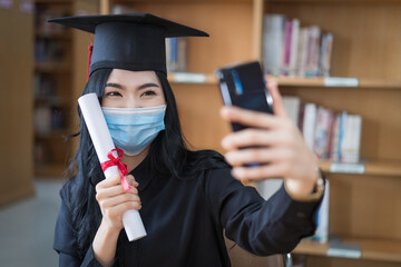 A young cheerful Asian woman university graduates in graduation gown and cap wears a face mask shows a degree certificate via video call to celebrate her education achievement on the commencement day