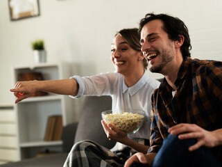 Wall Mural - Young loving couple relaxing and laughing on sofa together. Happy couple watching movie at home..