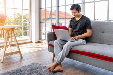 Happy young Asian man using laptop computer sitting o sofa in living room, work from home concept.