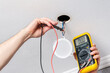 Electrician hands measure with tester power supply voltage before installing modern LED light bulb on the ceiling close-up. Energy saving technologies concept