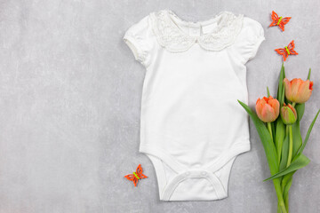 White baby girl bodysuit mockup on the gray concrete background with tulips flowers and spring decoration. Design onesie template, print presentation mock up. Top view. Flat Lay. Copy space.