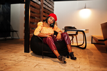 Wall Mural - Elegant afro american woman in red french beret, big gold neck chain polka dot blouse and leather pants sit on pouf.