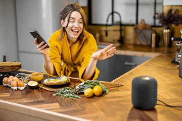 Happy woman in bathrobe controlling home devices with a voice commands, speaking to a smart column during breakfast time on the kitchen at home. Smart home concept