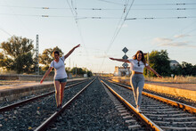 Carefree Female Friends With Arms Outstretched Walking On Railroad Track Against Sky At Sunset