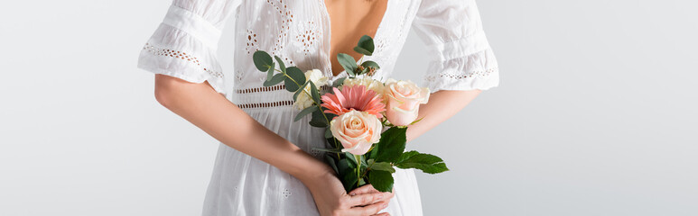 Sticker - cropped view of young woman in dress holding bouquet of spring flowers behind back isolated on white, banner