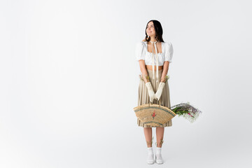 Sticker - full length of joyful young woman in spring outfit holding straw bag with flowers on white