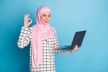 Wall Mural - Photo of young girl happy hold computer show okey sign choose recommend advert wear pink hijab isolated over blue color background