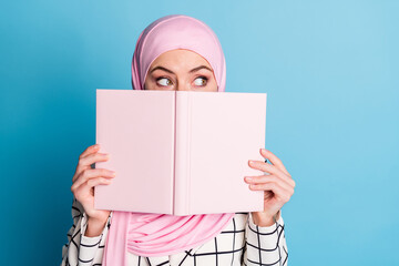 Wall Mural - Close-up portrait of funny curious muslimah girl closing face book looking aside isolated over vivid blue color background