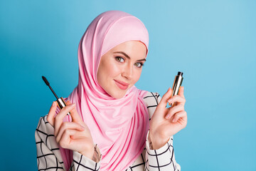 Wall Mural - Close-up portrait of pretty cheery feminine muslimah girl holding in hands new decorative mascara isolated over bright blue color background