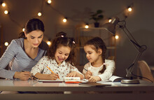 Young Mother With Little Daughters Drawing On Paper Doing Homework Sitting At Desk Table. Happy Family Having Fun Spending Evening Time Together In Modern Home Living Room Decorated With Lights