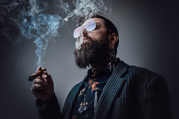 Elegant man in retro style poses in dark background smoking cigar. Portrait of a bearded guy with stylish coiffure.