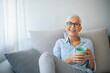 © Dragana Gordic - Delicious drink. Cheerful elderly woman sitting on the sofa in her living room and holding a cup of coffee while smiling at the camera. Happy beautiful dreamy old senior middle aged woman sit on sofa