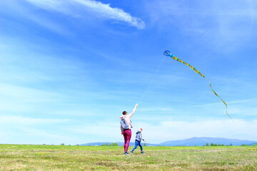 Happy family, mom and son, playing with flying kite on meadow in beautiful spring sunny day, blue sky in background