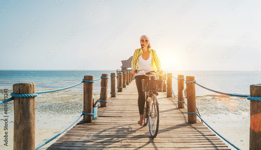 Portrait of a happy smiling woman dressed in light summer clothes and sunglasses riding a bicycle on the wooden sea pier and looking at camera. Careless vacation in tropical countries concept image - obrazy, fototapety, plakaty 