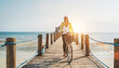 Portrait of a happy smiling woman dressed in light summer clothes and sunglasses riding a bicycle on the wooden sea pier and looking at camera. Careless vacation in tropical countries concept image