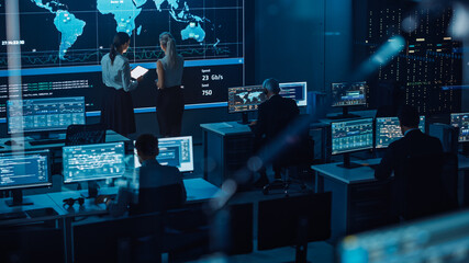 Poster - Two Software Engineers Talking Next to a Big Screen in a Modern Monitoring Office with Live Analysis Feed with Charts. Monitoring Room Big Data Scientists and Managers Sit in Front of Computers.