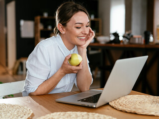 Happy woman using the laptop, having video call. Beautiful girl eating apple while talking with friends.