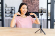 © Vadim Pastuh - Asian female blogger smiling, sitting at the desk, at home, talking in front of mobile phone standing on a tripod, giving her expert opinion, recommending, shows with her hands to an copy space