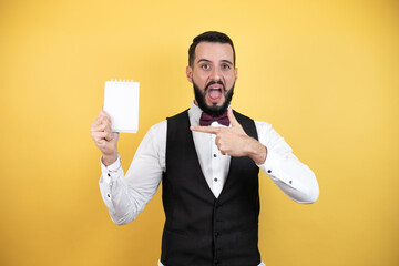 Wall Mural - Young man with beard wearing bow tie and vest smiling and showing blank notebook