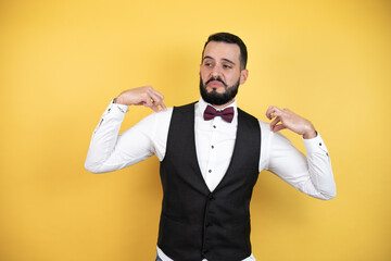 Wall Mural - Young man with beard wearing bow tie and vest holding his t-shirt with a successful expression casual clothes