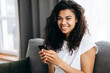 © Kateryna - Joyful attractive female sit on the sofa, using smartphone. African american woman chatting with friends or boyfriend, browsing internet, smiling and looks at the camera