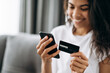 © Kateryna - A credit card and smartphone in female hands. Joyful african american young woman in defocus looks at the phone screen, shopping online. Credit card and phone in the focus