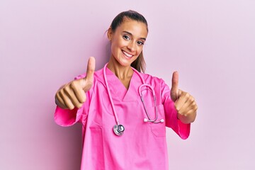 Wall Mural - Young hispanic woman wearing doctor uniform and stethoscope approving doing positive gesture with hand, thumbs up smiling and happy for success. winner gesture.