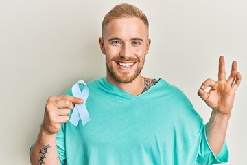 Wall Mural - Young caucasian man holding blue ribbon doing ok sign with fingers, smiling friendly gesturing excellent symbol