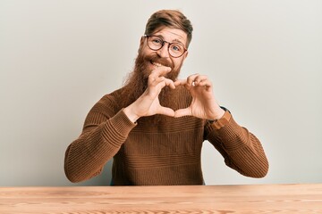 Canvas Print - Young irish redhead man wearing casual clothes and glasses sitting on the table smiling in love doing heart symbol shape with hands. romantic concept.