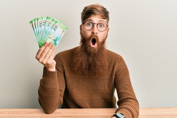 Poster - Young irish redhead man holding russian 200 ruble banknotes sitting on the table scared and amazed with open mouth for surprise, disbelief face