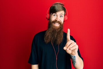 Canvas Print - Redhead man with long beard listening to music using headphones showing and pointing up with finger number one while smiling confident and happy.