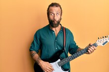 Handsome Man With Beard And Long Hair Playing Electric Guitar Making Fish Face With Mouth And Squinting Eyes, Crazy And Comical.