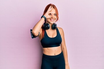 Canvas Print - Young beautiful redhead woman wearing gym clothes and using headphones doing ok gesture with hand smiling, eye looking through fingers with happy face.