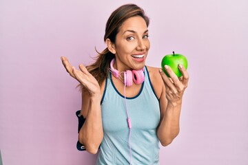 Young latin woman wearing gym clothes, using headphones and eating green apple celebrating achievement with happy smile and winner expression with raised hand