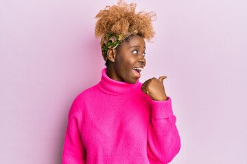 Canvas Print - Young african woman with afro hair wearing casual winter sweater pointing thumb up to the side smiling happy with open mouth