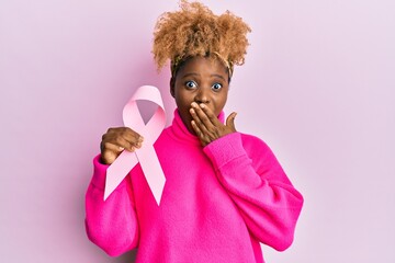 Canvas Print - Young african woman with afro hair holding pink cancer ribbon covering mouth with hand, shocked and afraid for mistake. surprised expression