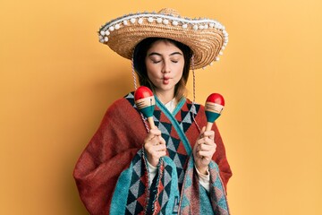 Wall Mural - Young caucasian woman holding mexican hat using maracas making fish face with mouth and squinting eyes, crazy and comical.