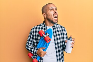 Canvas Print - Hispanic adult skater man holding skate and graffiti spray angry and mad screaming frustrated and furious, shouting with anger looking up.