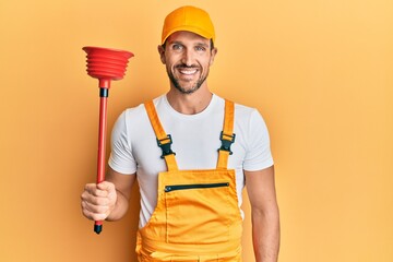 Wall Mural - Young handsome man wearing plumber uniform holding toilet plunger looking positive and happy standing and smiling with a confident smile showing teeth