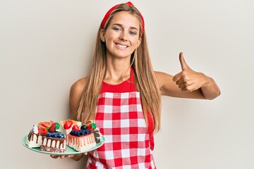 Young blonde woman wearing baker uniform holding homemade cakes smiling happy and positive, thumb up doing excellent and approval sign